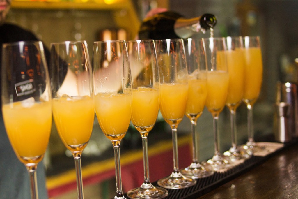 A row of champagne flutes filled with a vibrant orange beverage, possibly mimosa or a similar cocktail, with a bartender pouring in the background.