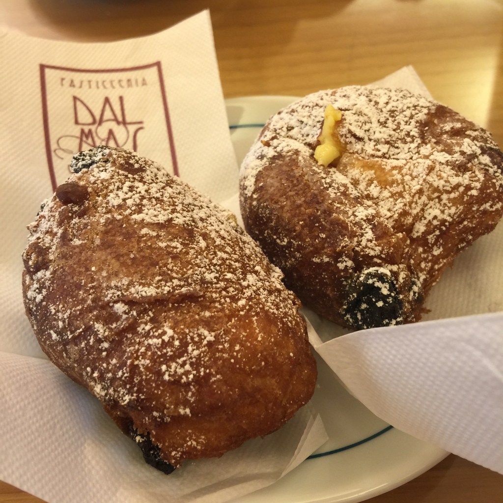 Two fritelle pastries dusted with powdered sugar on a plate, accompanied by a napkin featuring the Dal Mas logo.