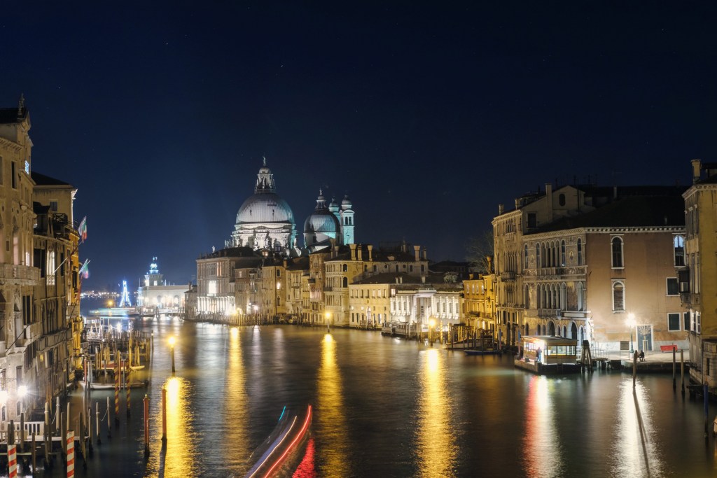 Night view of the Grand Canal in Venice, illuminated by street lamps and reflecting the historic buildings and domes under a starry sky.