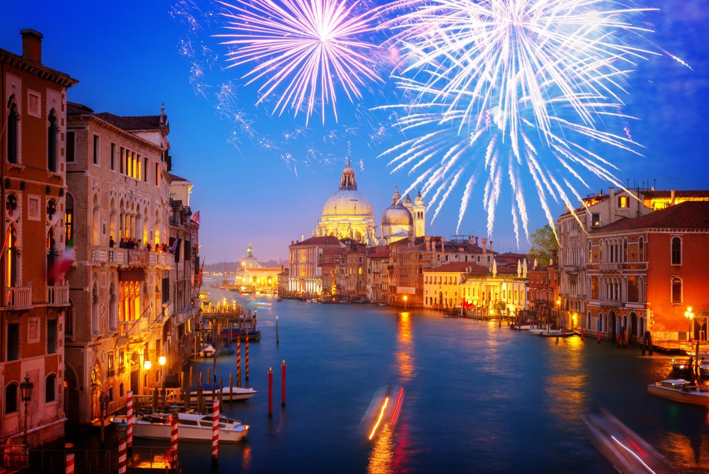 Fireworks illuminate the night sky over the Grand Canal in Venice, with historic buildings reflecting in the water, creating a festive atmosphere for New Year's Eve.