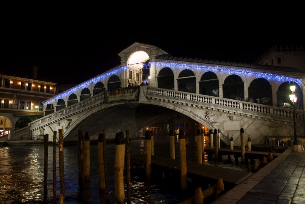 The Rialto Bridge in Venice, illuminated at night with blue lights, spanning across the Grand Canal, surrounded by water and historic buildings.