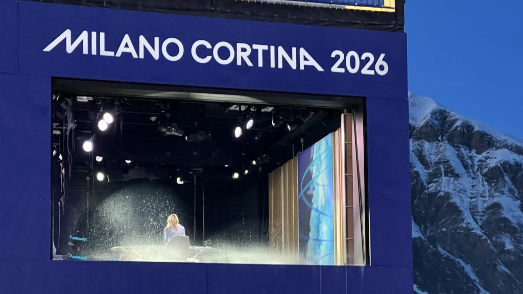 Stage setup for Milano Cortina 2026 with a performer in front of bright lights and a backdrop featuring snow-covered mountains.