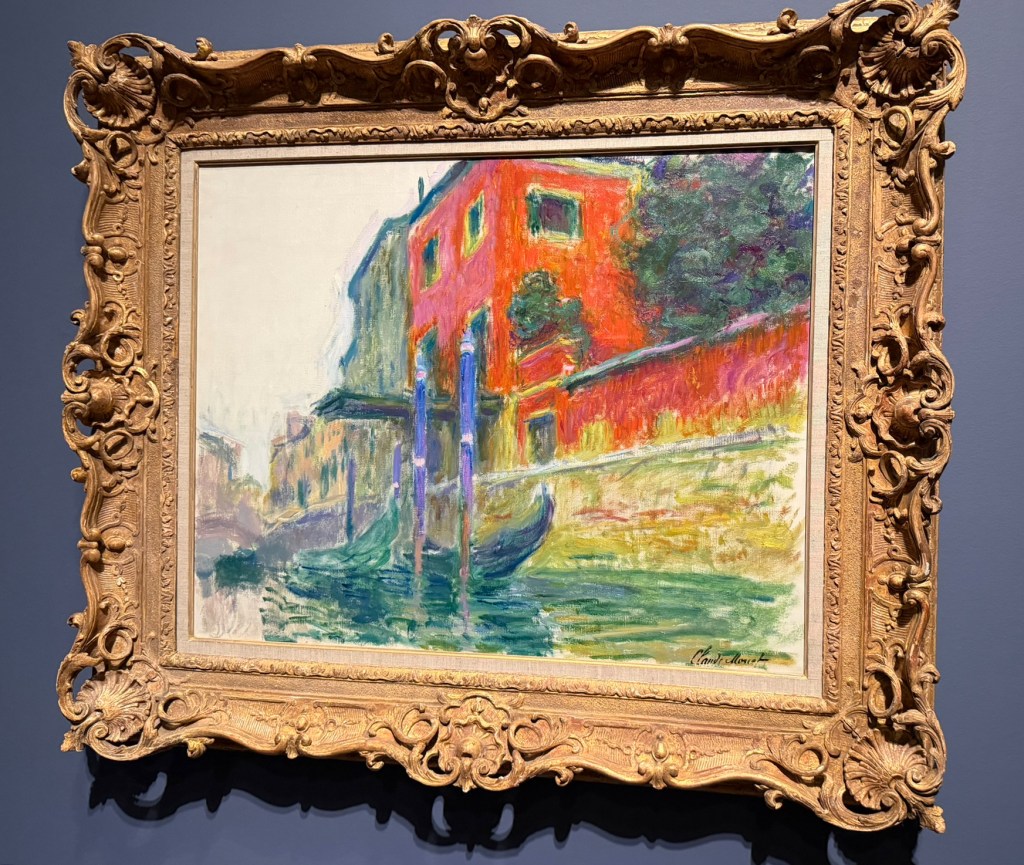 A colorful impressionist painting depicting a canal scene with vibrant red buildings and blue poles, framed in an ornate gold frame.